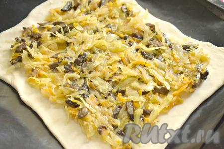 Один пласт размороженного слоёного теста выкладываем на стол, присыпанный мукой. Немного раскатываем тесто и выкладываем на противень, застеленный ковриком (или пергаментом) для выпечки. Поверх теста, отступив от краёв 1,5-2 сантиметра, равномерно выкладываем остывшую начинку из капусты и грибов.