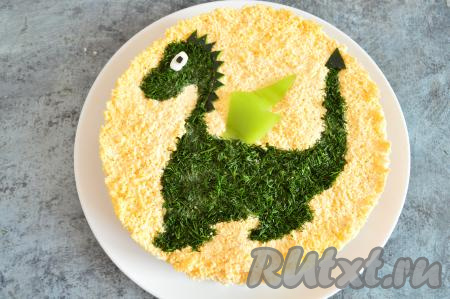 Снять аккуратно трафарет и украсить салат. Из отложенного белка вырезать круглый или овальный глаз, выложить поверх укропа. Поверх белка выложить зрачок, вырезанный из маслины (или сделать из горошины чёрного перца). Крыло вырезать из болгарского перца, выложить рядом со спиной дракона. Из огурца вырезать треугольный кончик хвоста и шипы на голове, приложить их в нужное место фигурки. Снять формовочное кольцо, отправить салат пропитаться в холодильник хотя бы минут на 30, а затем можно подавать к столу.