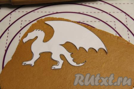 Охладившееся пряничное тесто на слегка присыпанной мукой поверхности раскатать скалкой в пласт толщиной примерно 5 миллиметров. На тесто выложить трафарет дракона (или вырубку для пряников в виде дракона) и вырезать пряники ножом по контуру трафарета. Я заранее подготовила бумажный трафарет и с помощью него вырезала из теста драконов. Бумажный трафарет в домашних условиях сделать несложно, нужно лишь найти в Интернете подходящую картинку, распечатать и вырезать.