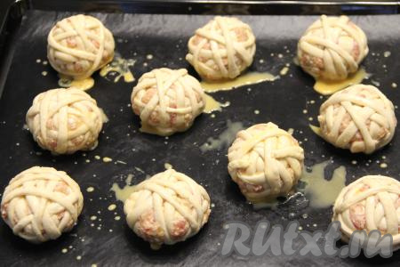 Выложить получившиеся мясные клубочки в слоёном тесте на противень, предварительно застеленный тефлоновым ковриком (или пергаментом). Яичный желток взбить в небольшой ёмкости вилкой. Смазать клубочки взбитым желтком и, по желанию, присыпать кунжутом.