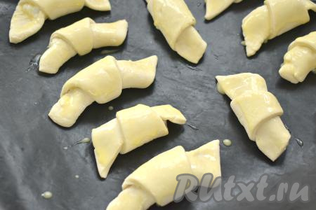 На противень, застеленный ковриком (или пергаментом) для выпечки выкладываем рогалики с творогом "хвостиком" вниз. Отложенный ранее желток перемешиваем до однородности с 3 столовыми ложками воды, получившейся смесью смазываем рогалики.