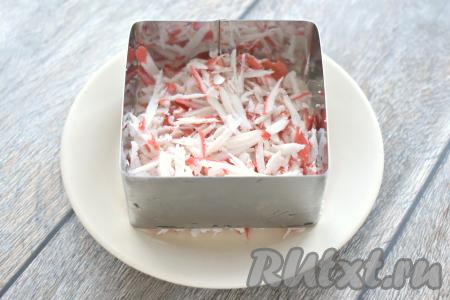 Заранее отвариваем до готовности морковь в кожуре и яйца, затем даём им остыть и очищаем. Крабовые палочки, если нужно, размораживаем, достаём из упаковки, натираем на крупной тёрке. На плоскую тарелку ставим сервировочную форму. Я раскладывала ингредиенты по 2 сервировочным квадратным формам со стороной 9 сантиметров, формируя 2 порции салата. Первым слоем в сервировочную форму выкладываем половину натёртых крабовых палочек. Вторую половину натёртых палочек кладём во вторую форму. 