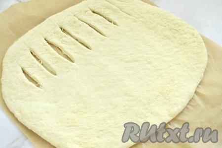Поднявшееся дрожжевое тесто обминаем руками. На столе, присыпанном мукой, раскатываем дрожжевое тесто в прямоугольный пласт толщиной примерно 7-9 миллиметра. С одной стороны пласта делаем надрезы ножом, оставляя края нетронутыми, как на фото.