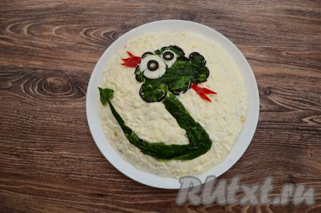 Теперь можно приступать к оформлению новогоднего салата. Для этого из маслин, нарезанных на тонкие полоски, нужно сформировать контур головы. Середину контура заполнить ранее отложенными кусочками кожуры огурца. Белки глаз вырезать из яичных белков, выложить поверх кожуры. Зрачки сделать из маслин. Тело и хвост змеи будут зелёными, выложить их из кожуры огурца. Корону и язык вырезать из болгарского перца и разместить на салат.