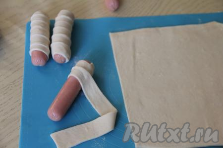 Каждую сосиску обернуть в полоску теста внахлёст, слегка растягивая тесто. После того как вся сосиска будет обёрнута в тесто, край полоски нужно закрепить. Если длины полоски теста не хватает, чтобы обернуть всю сосиску, тогда возьмите следующую полоску теста и продолжите оборачивать сосиску в тесто внахлёст. После того как вся сосиска будет обёрнута в тесто, лишнее тесто обрежьте и используйте оставшийся кусочек полоски теста для оборачивания следующей сосиски.