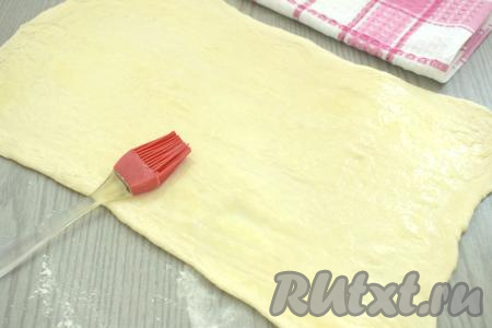 Прежде всего достаём слоёное тесто из морозилки, оставляем его размораживаться при комнатной температуре (ориентировочно на 30-40 минут, тесто должно стать достаточно мягким). Для этой выпечки можно взять слоёное тесто как дрожжевое (я его и купила), так и бездрожжевое. На присыпанной мукой поверхности раскатываем слоёное тесто в достаточно тонкий прямоугольник (у меня получился прямоугольник толщиной 3 миллиметра, размером 22 сантиметра на 35 сантиметров). В небольшой ёмкости взбиваем вилкой до однородности сырое яйцо. Поверхность раскатанного теста смазываем взбитым яйцом. Немного яйца оставьте для смазывания полосок перед выпечкой.