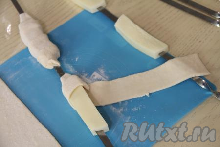 Каждый брусок сыра обернуть в полоску из слоёного теста, накладывая тесто немного внахлёст и слегка растягивая его в процессе обматывания сыра. Слоёное тесто должно закрывать сыр с двух сторон. Тесто нужно хорошо прижать к шампурам с двух сторон, чтобы сыр был хорошо закрыт и не вытекал во время приготовления на мангале.
