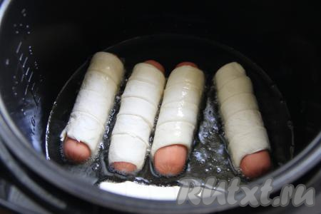 В чашу мультиварки влить растительное масло, выставить режим "Жарка". Как только масло прогреется, выложить в чашу в один слой сосиски, завёрнутые в слоёное тесто.