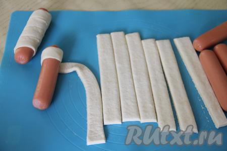 Каждую сосиску завернуть в полоску из слоёного теста, накладывая тесто внахлёст и слегка растягивая полоску из теста во время оборачивания. Конец полоски теста хорошо закрепить.