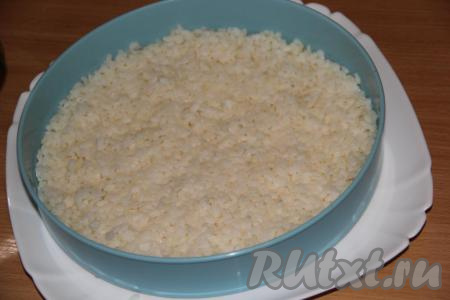 Взять плоскую тарелку. На неё установить бортик для салата, можно использовать кольцо от разъёмной формы для тортов. Выложить половину сваренного риса на тарелку и утрамбовать ложкой.