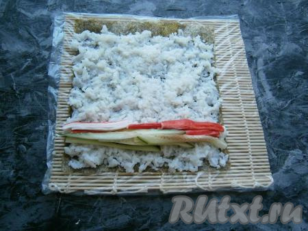 Ближе к нижнему краю листа горизонтально выложить крабовые палочки, разрезанные на тонкие полоски, рядом расположить огурцы, нарезанные на такие же полосы (как на фото).