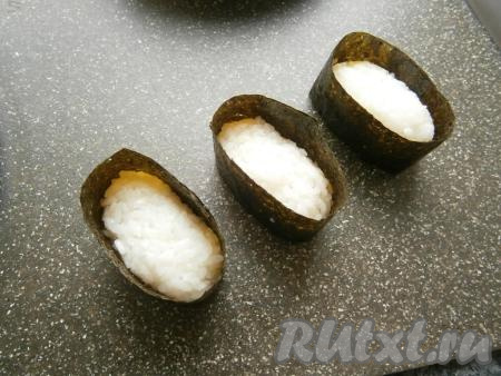 Лист нори нарезать на прямоугольники, рассчитав высоту так, чтобы лист был выше брусочков риса на 1-2 см. Рисовые брусочки обернуть нарезанными листами нори, конец склеить водой (или рисинкой). Получатся такие своеобразные "чашечки".