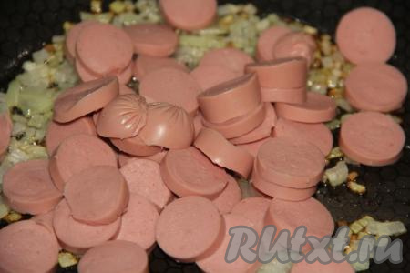 В сковороду влить масло, поставить на средний огонь, выложить лук и обжарить его до золотистого цвета, периодически помешивая. Когда лук станет золотистым, добавить на сковороду нарезанные сосиски и чеснок.