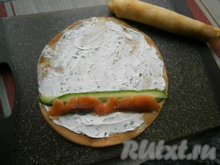 Каждый остывший блин полностью смазать слоем творожного сыра. На нижний край блина выложить нарезанную тонкими пластинами красную рыбу и нарезанный тонкими слайсами с помощью овощечистки свежий огурчик.