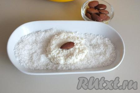 Подготовьте миндаль. Важно, чтобы он не был горьким. В тарелочку насыпьте кокосовую стружку. Зачерпните столовой ложкой творожную массу, выложите её на кокосовую стружку. Немного прижмите творожную массу, чтобы получилась лепёшка. На серединку творожной "лепёшки" положите миндаль.