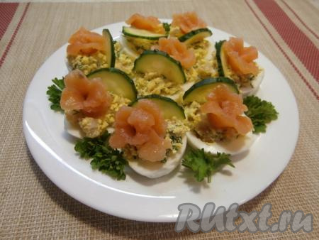 Оставшуюся красную рыбу нарезать на тонкие полоски, свернуть в виде розочек и украсить фаршированные яйца сверху. Рядом разместить по половинке кружочка свежего огурца. Украсить закуску зеленью петрушки.