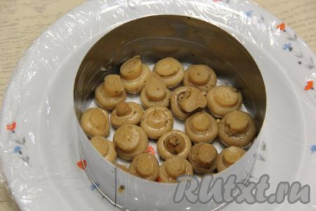 Этот салат формируется слоями. Удобно формировать его в кольце для формирования салатов или в кольце от разъёмной формы для выпечки бисквитов. Я формировала 2 салата в двух кулинарных кольцах диаметром 12 см, поэтому равномерно распределяла ингредиенты по двум кольцам. Итак, плоскую тарелку нужно застелить пищевой плёнкой (плёнка поможет перевернуть этот салат-перевёртыш после его формирования). Первым слоем внутрь кольца выложить целые шампиньоны шляпками вниз. 