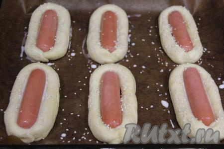 Сосиску обернуть жгутиком теста, скрепить края. Выложить булочки с сосисками на противень, застеленный тефлоновым ковриком (если нет такого коврика, можно застелить пергаментом), и оставить в тёплом месте на 30 минут, накрыв полотенцем. Затем смазать тесто молоком (или водой) и, по желанию, присыпать кунжутом.