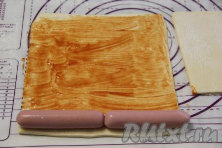 Немного присыпать пласт теста мукой и слегка раскатать скалкой. Я разделила тесто на два пласта и раскатала каждый пласт под размер длины двух сосисок. Смазать каждый раскатанный пласт теста кетчупом и на край пласта выложить сосиски, очищенные от плёнки.