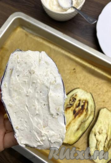 Достать пластины баклажанов из духовки, дать им немного остыть. В это время нарезать помидор на тонкие ломтики. Когда запечённые баклажаны остынут, смазать каждую полоску с одной стороны смесью творожного сыра и чеснока.