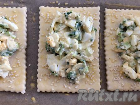 В небольшой ёмкости яичный желток смешать с 2 чайными ложками молока. Свободные края слоек смазать при помощи силиконовой кисточки смесью желтка и молока, затем посыпать кунжутом. Отправить противень в разогретую духовку и выпекать слойки с яйцом и зелёным луком минут 20 (до подрумянивания теста) при температуре 180 градусов.