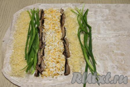 Начинку на лаваш нужно будет выкладывать полосками, начиная от узкого края лаваша и отступая от краёв лаваша по 1,5 сантиметра. Сначала выложить в виде полоски половину натёртого сыра, как на фото. Рядом выложить половину зелёного лука. Затем сформировать полоску из шпрот (я выложила шпротинки в 2 ряда). Следующей полоской будут натёртые яйца. Рядом расположить полоску из оставшихся шпрот. Затем сформировать полоску из оставшегося натёртого сыра и полоску из зелёного лука.