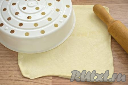 Слегка присыпаем стол мукой, выкладываем размороженный пласт теста и раскатываем его. Раскатанное тесто должно быть по размеру чуть больше диаметра съёмной чаши мультиварки. Нам нужно вырезать круг. Для того чтобы определить диаметр круга, который нам нужен, я приложила к раскатанному тесту поддон от мультиварки для варки на пару (у меня мультиварка "Редмонд") и вырезала круг нужного диаметра. Тесто, оставшееся после вырезания круга, можно собрать в комок, раскатать в лепёшку и приготовить ещё одну маленькую пиццу.