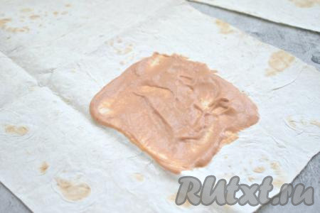 Тонкий лаваш разрезаем на 2 равные части. Примерный размер одной части лаваша - 23 сантиметра на 30 сантиметров. Всего у меня из 2 листов лаваша получилось 4 заготовки. В небольшой ёмкости смешиваем томатный соус (или кетчуп) и майонез. Одну заготовку лаваша раскладываем на столе. Ближе к узкому краю, равномерно отступив от краёв, немного смазываем лаваш частью смеси томатного соуса и майонеза, как на фото.