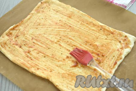 Пицца, как правило, бывает круглой формы, но я решила сделать её прямоугольной, так как слоёное тесто продаётся в прямоугольных пластах. На посыпанный мукой стол выкладываем пласт теста, раскатываем его до толщины примерно 3 миллиметра. У меня получился прямоугольник из теста размером 29 сантиметров на 20 сантиметров. Раскатанное тесто помещаем сразу на противень, застеленный бумагой (или ковриком) для выпечки. Смазываем раскатанное тесто томатным соусом (или кетчупом).