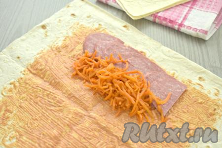 Копчёную колбасу нарезаем на полукружочки. На одну сторону лаваша, отступая от краёв лаваша 5-6 сантиметров, выкладываем внахлёст в один ряд половину нарезанной колбасы. Корейскую морковку отжимаем от лишней жидкости. Если полоски моркови длинные, разрезаем их на 3-4 части. Рядом с колбасой выкладываем половину корейской морковки.