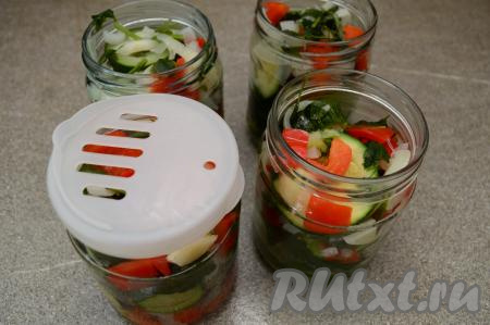 После того как овощи с момента закипания воды проварятся 2 минуты, выключить огонь. Равномерно разложить в кастрюлю по 4 банкам кабачки, болгарский перец, лук, чеснок и зелень. После того как овощи будут равномерно распределены по банкам, нужно жидкость, которая попала в банки вместе с овощами, слить обратно в кастрюлю, в которой варились кабачки с перцем. Очень удобно сливать жидкость из банок, надев на баночку специальную крышку с отверстиями. Банки с овощами прикрыть крышками.