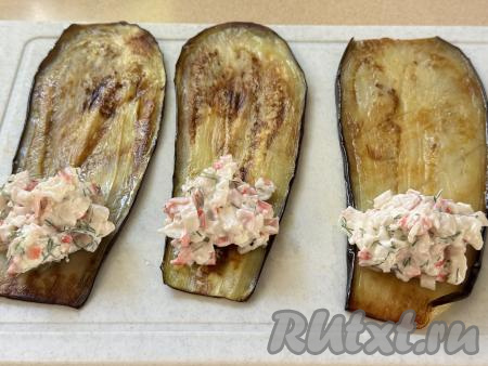 Выложить полоски баклажана на разделочную доску. Положить на более узкий край каждой полоски из баклажана по 1 чайной ложке начинки из сыра и крабовых палочек.