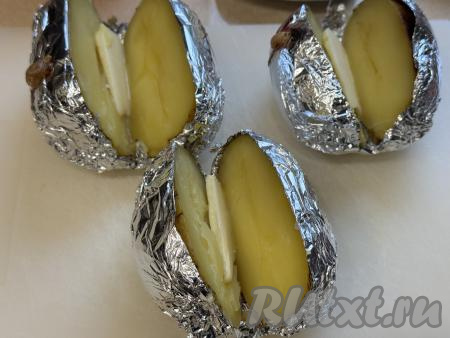 Готовый запечённый картофель переложить на доску. Не снимая фольгу, разрезать каждую картошину на 2 части в длину, не прорезая до конца. Вложить внутрь каждой горячей картошины кусочек сливочного масла.&nbsp;