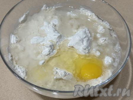 Добавить яйцо, растительное масло и 150 миллилитров воды, вначале перемешивать тесто столовой ложкой. А после того как перемешивать тесто ложкой станет тяжело, продолжить замешивать его руками.