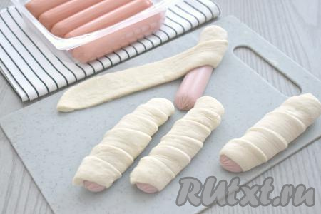 Сосиски разрезаем вдоль на две половинки. Каждую половину сосиски оборачиваем внахлёст в полоску из дрожжевого теста, немного растягивая в процессе заворачивания полоску теста руками. Накрываем сформированные сосиски в тесте полотенцем и оставляем на расстойку минут на 15.
