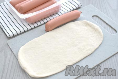Поднявшееся дрожжевое тесто обминаем и делим на 3 равных части. На слегка присыпанной мукой поверхности вначале одну часть теста раскатываем в прямоугольную заготовку по длине в два раза больше, чем длина сосиски. Ширина прямоугольной заготовки у меня получилась 12 сантиметров.