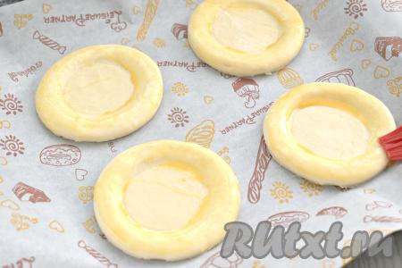 На противень, застеленный пергаментом (или ковриком) для выпечки, выкладываем заготовки из теста для ватрушек. В небольшой ёмкости вилкой взбиваем желток до однородности. С помощью силиконовой кисточки смазываем бортики ватрушек взбитым желтком.&nbsp;
