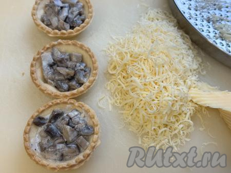 Разложить грибную начинку по песочным тарталеткам. Сыр натереть на мелкой тёрке. Я разложила начинку по 10 тарталеткам, но количество тарталеток, которые вы заполните, зависит прежде всего от размера тарталеток, которые вы используете.
