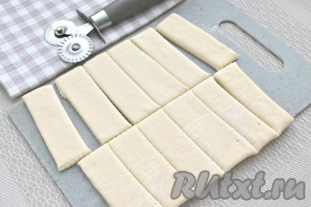 Поверхность стола (или разделочной доски) посыпаем немного мукой, выкладываем размороженный пласт слоёного теста. Не раскатывая тесто, с помощью пиццерезки (или острого ножа) нарезаем его на прямоугольные полоски размером примерно 3 сантиметра на 7,5 сантиметров. В результате у меня получились 12 полосок.