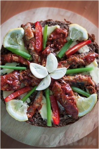 На верх торта выложите по кругу кильку, перышки зеленого лука, нарезанный тонкими полосками сладкий перец, дольки лимона. В центре разложите дольки чеснока в виде цветочка.