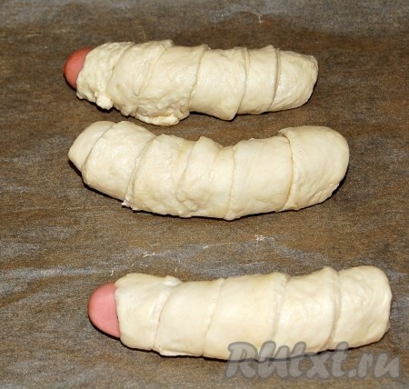 Накручивать полоску теста на сосиску, внахлест. Выложить сосиски в тесте на противень, застеленный бумагой для выпечки.