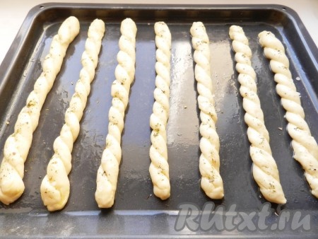 Вначале один кусочек теста скатать в длинный тонкий жгутик, затем сложить его пополам и переплести, формируя хлебную палочку. Аналогично сформировать все хлебные палочки и выложить их на противень, смазанный маслом. Каждую палочку смазать оливковым маслом, посыпать итальянскими травами и крупной солью. Оставить ещё на 15-20 минут.