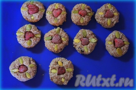 Духовку разогрейте до 180 градусов. Противень застелите бумагой для выпечки или силиконовым ковриком. Слегка влажными руками возьмите немного теста, скатайте небольшой шарик, слегка придавите его рукой и выложите на противень. В центр положите ягодку малины, украсьте печенье тыквенными семечками.
