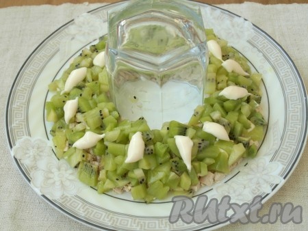 Следует сразу сварить куриное филе, морковь и яйца. Куриное филе нарезать кубиками. Также нарезать 2 киви, а 2 киви оставить для украшения. На плоскую тарелку в центр поставить стакан и выложить вокруг куриное филе. Сверху выложить кубики киви. В майонез выдавить чеснок и нанести его сверху киви.
