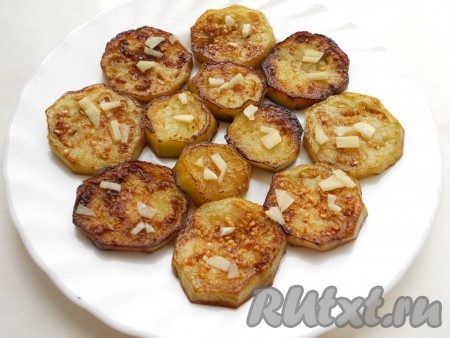 Из болгарских перцев удалить семена и плодоножку. Помидоры, перцы и зелень вымыть. Нарезать помидоры и перцы соломкой. Сыр натереть на мелкой тёрке. Зелень и зубчики чеснока мелко нарезать. На плоскую тарелку в один слой разложить половину обжаренных баклажанов, сверху посыпать половиной нарезанного чеснока, посолить.