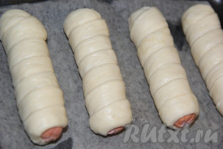 Противень для выпекания застелить пергаментом. Выложить сосиски в тесте на противень и оставить на 15-20 минут для подъёма.