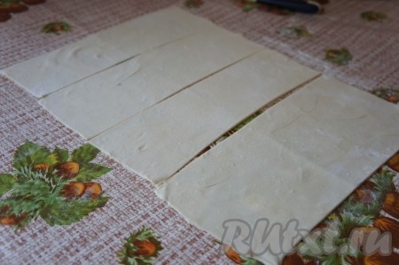 Слоёное тесто заранее достать из морозилки и разморозить при комнатной температуре. Стол присыпать мукой, раскатать слоёное тесто до толщины примерно 2 миллиметра. Разрезать раскатанное тесто на одинаковые прямоугольники (я разрезала на 4 прямоугольника), затем каждый прямоугольник разрезать по середине, начиная с широкой стороны, ещё на 2 части. У нас получатся 8 небольших прямоугольников из слоёного теста для формирования слоек.