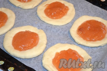 Переложить заготовки ватрушек на противень, застеленный бумагой для выпечки, на небольшом расстоянии друг от друга. Сделать в центре каждой заготовки углубление и заполнить его повидлом (на каждую заготовку я обычно выкладываю 2-3 чайных ложки яблочного повидла). Затем вилкой в небольшой ёмкости перемешать до однородности сырое яйцо. При помощи кулинарной кисти смазать яйцом края ватрушек. Поместить противень в заранее разогретую до 200-220 градусов духовку. Выпекать ватрушки с яблочным повидлом до золотистого цвета дрожжевого теста (примерно 20 минут).