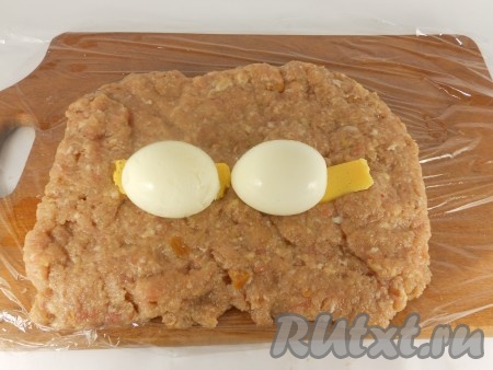 На пищевую плёнку выложить подготовленный фарш в форме прямоугольника. По середине, по желанию, разместить кусочки твёрдого сыра, затем в центре выложить 2 очищенных варёных яйца.