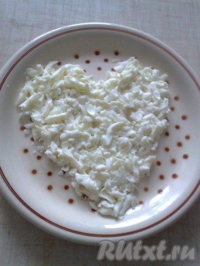 Лук для салата "Мимоза" должен быть как можно более нежным, поэтому очищенный лук нужно мелко нарезать, переложить в миску, залить кипятком, а после остывания воды лишнюю жидкость нужно полностью слить. Яйца заранее сварить вкрутую (в течение 10 минут после закипания), а затем остудить их полностью и очистить. На плоское блюдо начинаем слоями выкладывать ингредиенты в форме сердца. 1 слой состоит из 3 белков, натёртых на крупной тёрке.